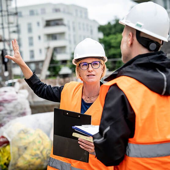 Eine Frau und ein Mann in Arbeitsschutzkleidung stehen auf einer Baustelle. Die Frau erklärt dem Mann etwas und unterstützt ihre Worte durch eine Handbewegung mit der linken Hand.