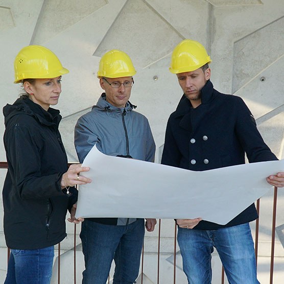 Drei Personen mit Helmen auf stehen vor einem Gebäude und betrachten gemeinsam eine große Karte.
