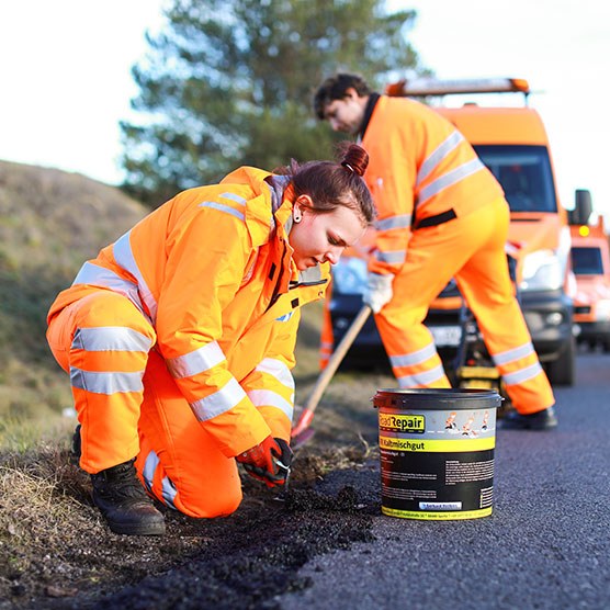 Zwei Mitarbeitende des Landesbetriebs Straßenwesen reparieren den Straßenrand mit Reparaturmischgut. Sie tragen dabei orange leuchtende Arbeitskleidung.